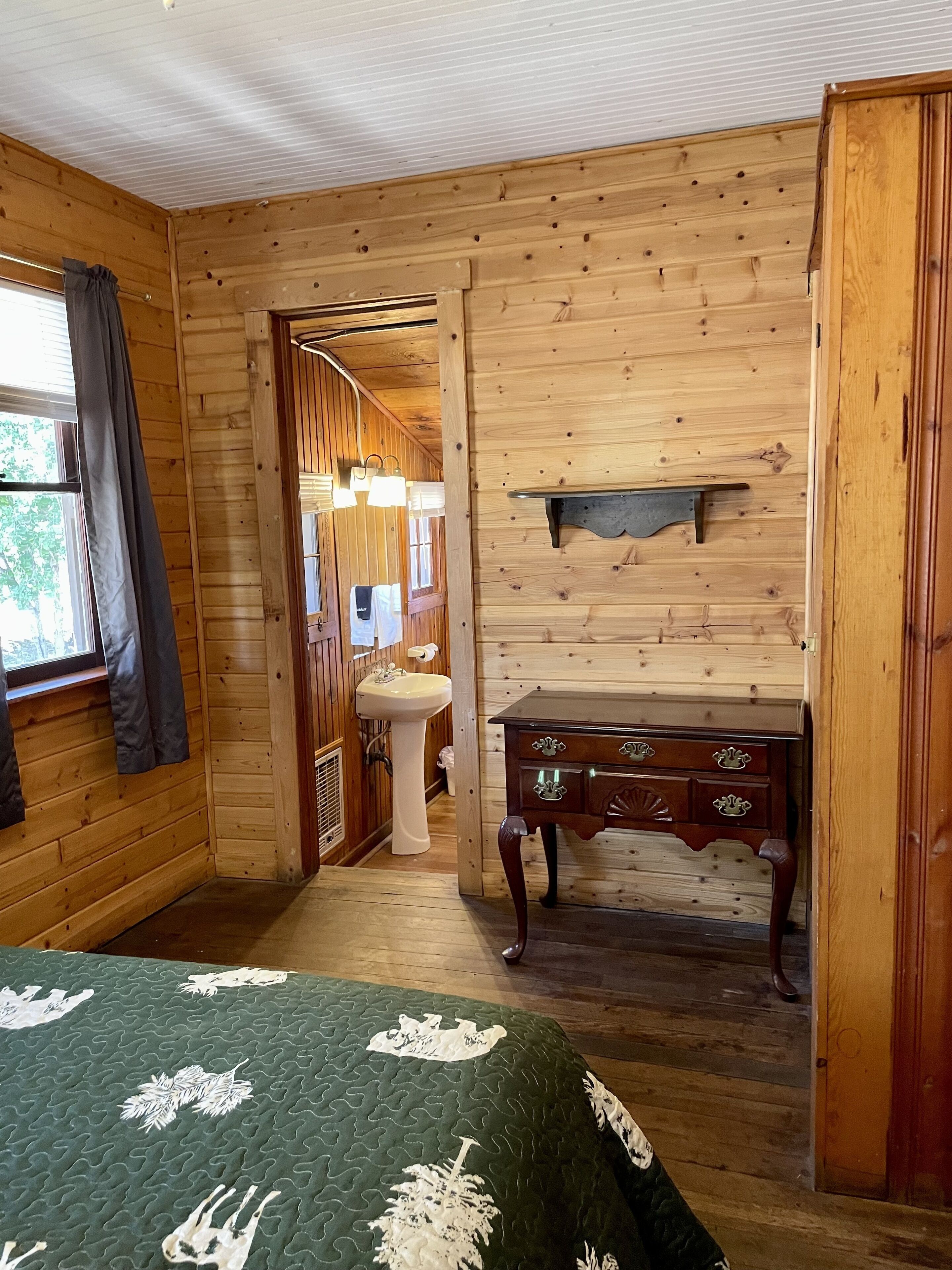 Quarto de cabana de madeira com cama, cómoda, pia e janela com cortinas.