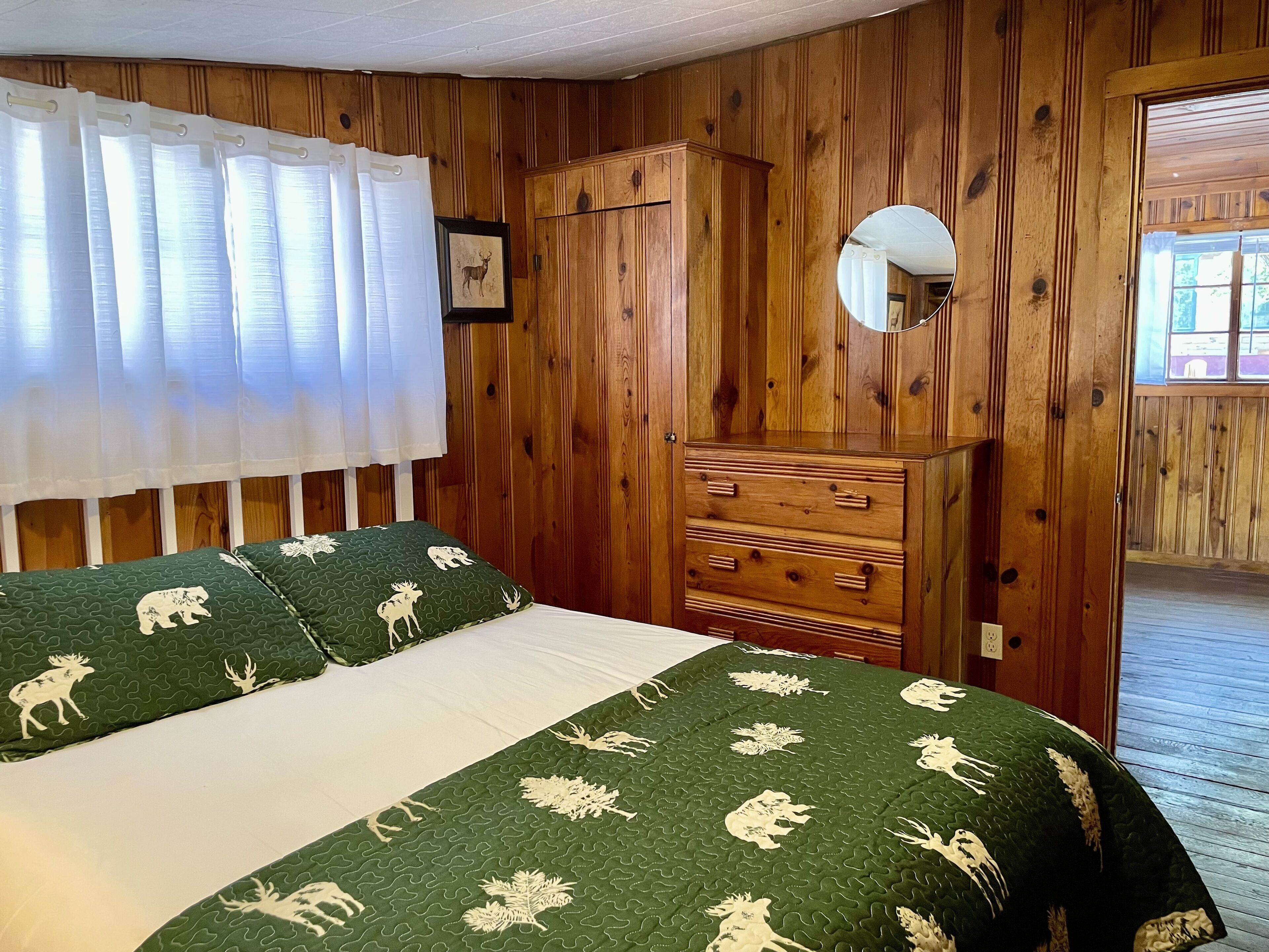 A room with wooden paneling, a bed with green bedding featuring animal prints, a wooden dresser, and a mirror on the wall.