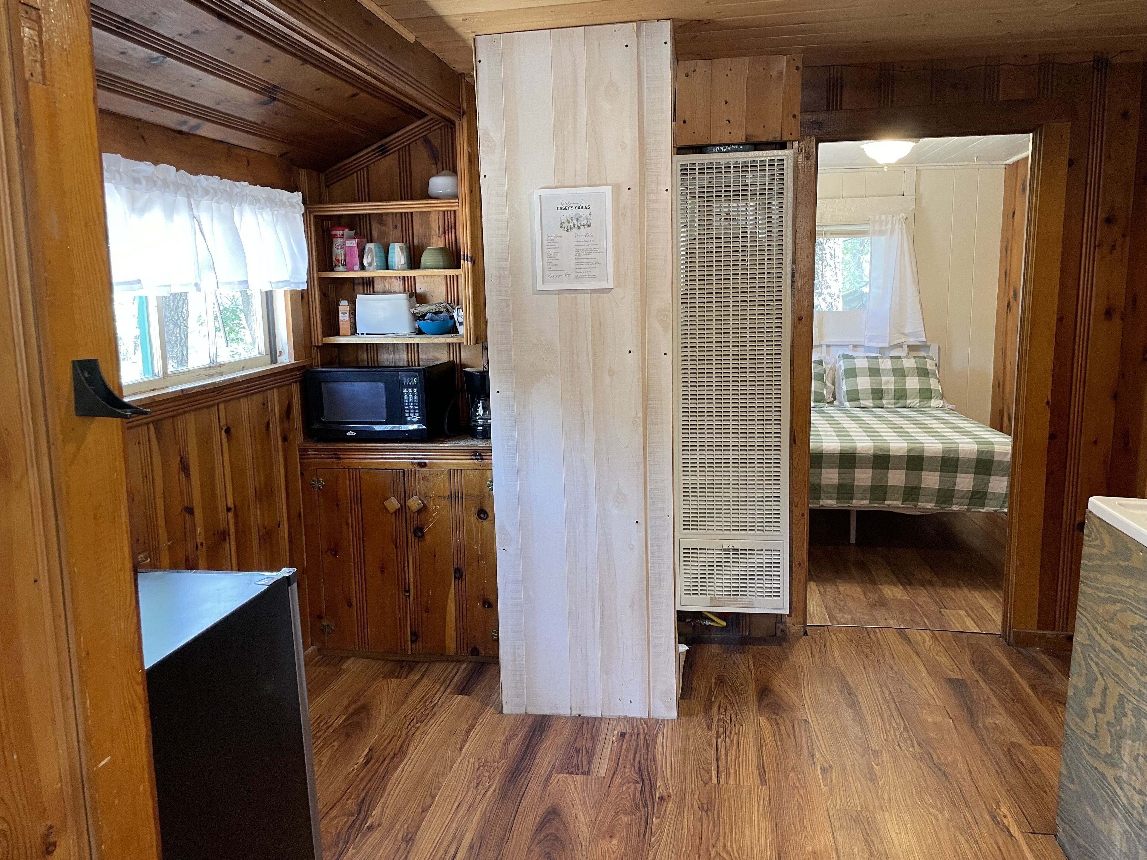 A cabin interior with a kitchen area featuring a microwave, shelves with various items, and a bed with a checkered bedspread in the adjacent room.