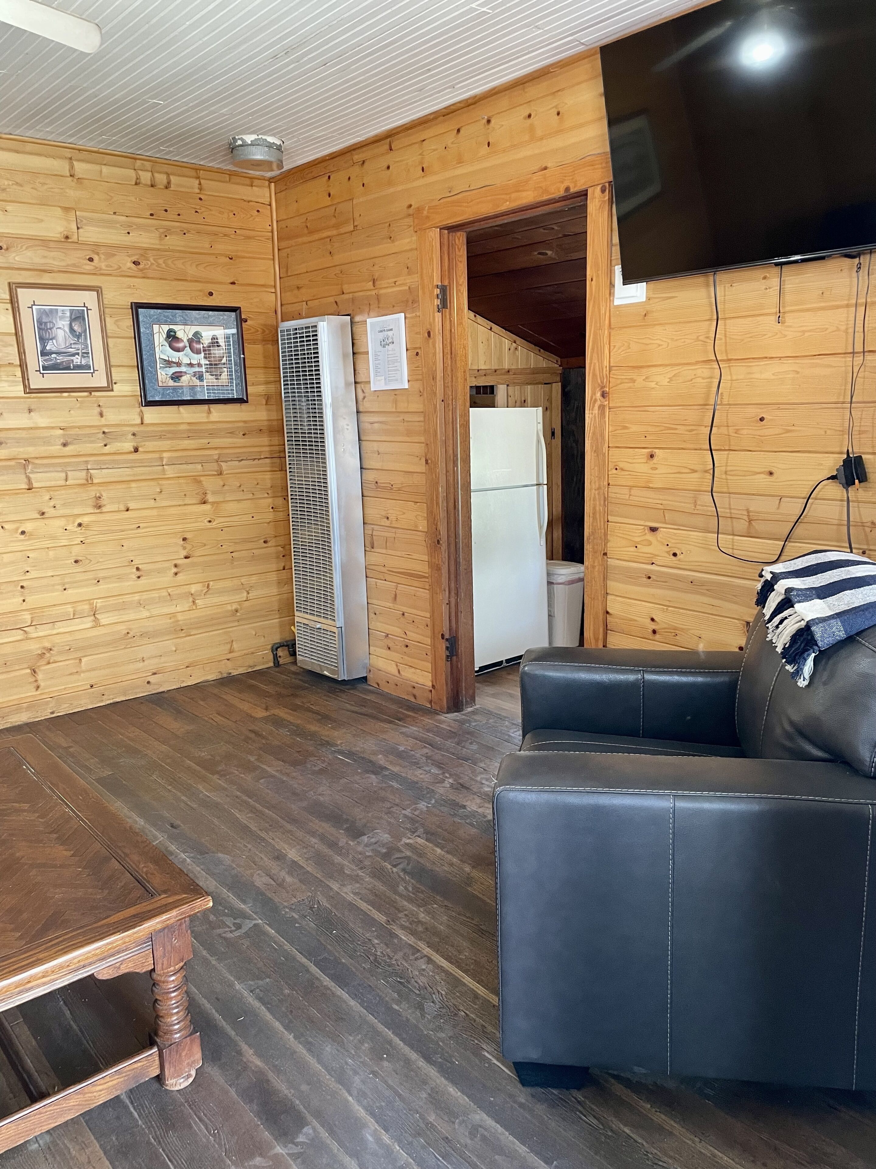 A wooden-paneled room with a black leather sofa, a wooden table, a white refrigerator, and a wall-mounted TV.