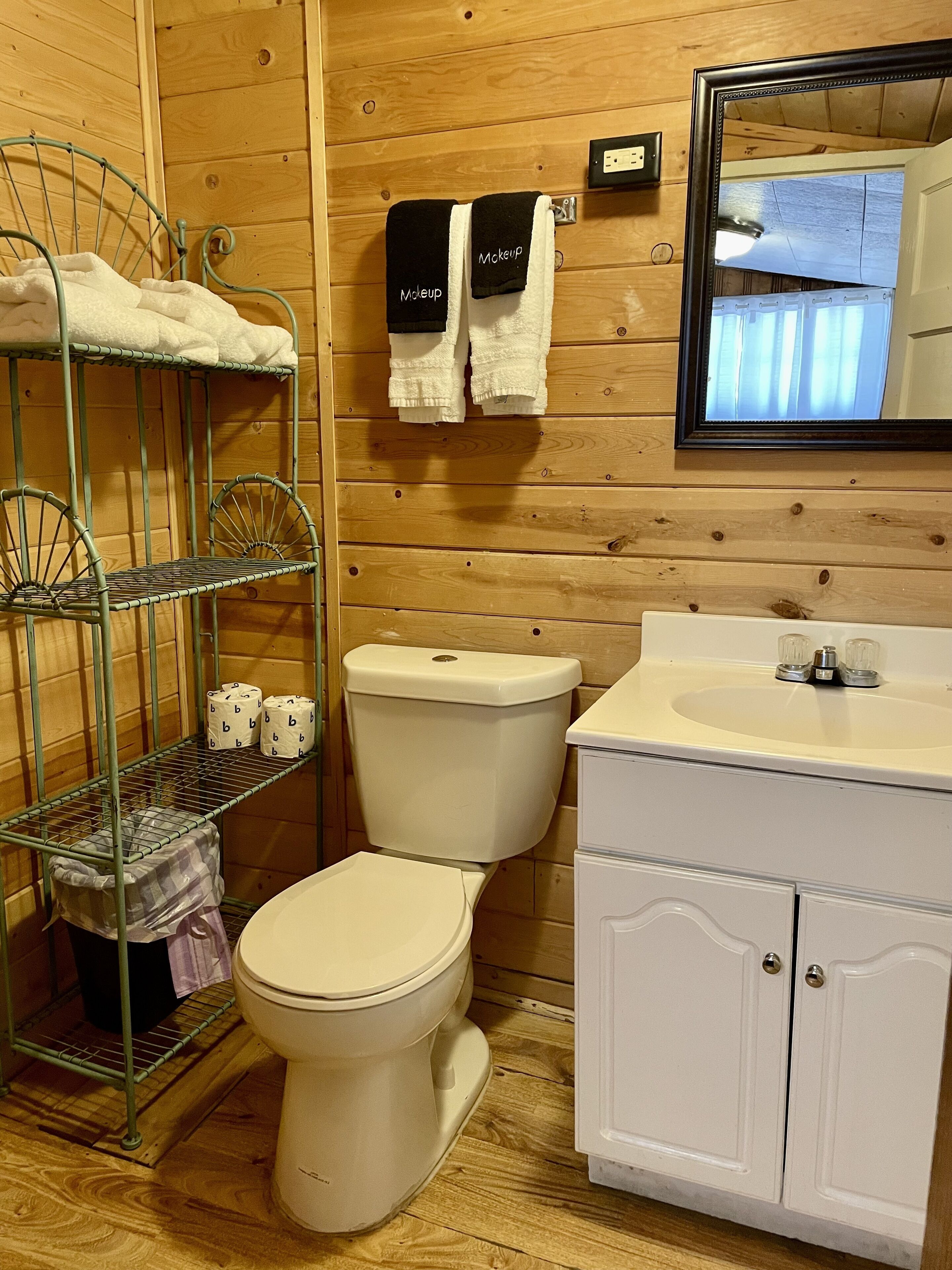 Un baño con paneles de madera, un inodoro blanco, una repisa metálica con toallas y un lavabo blanco con un espejo encima.