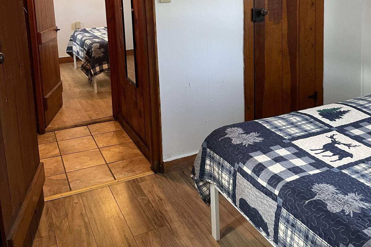A bedroom with a wooden door, a bed with a checkered quilt, and a hallway leading to another room.