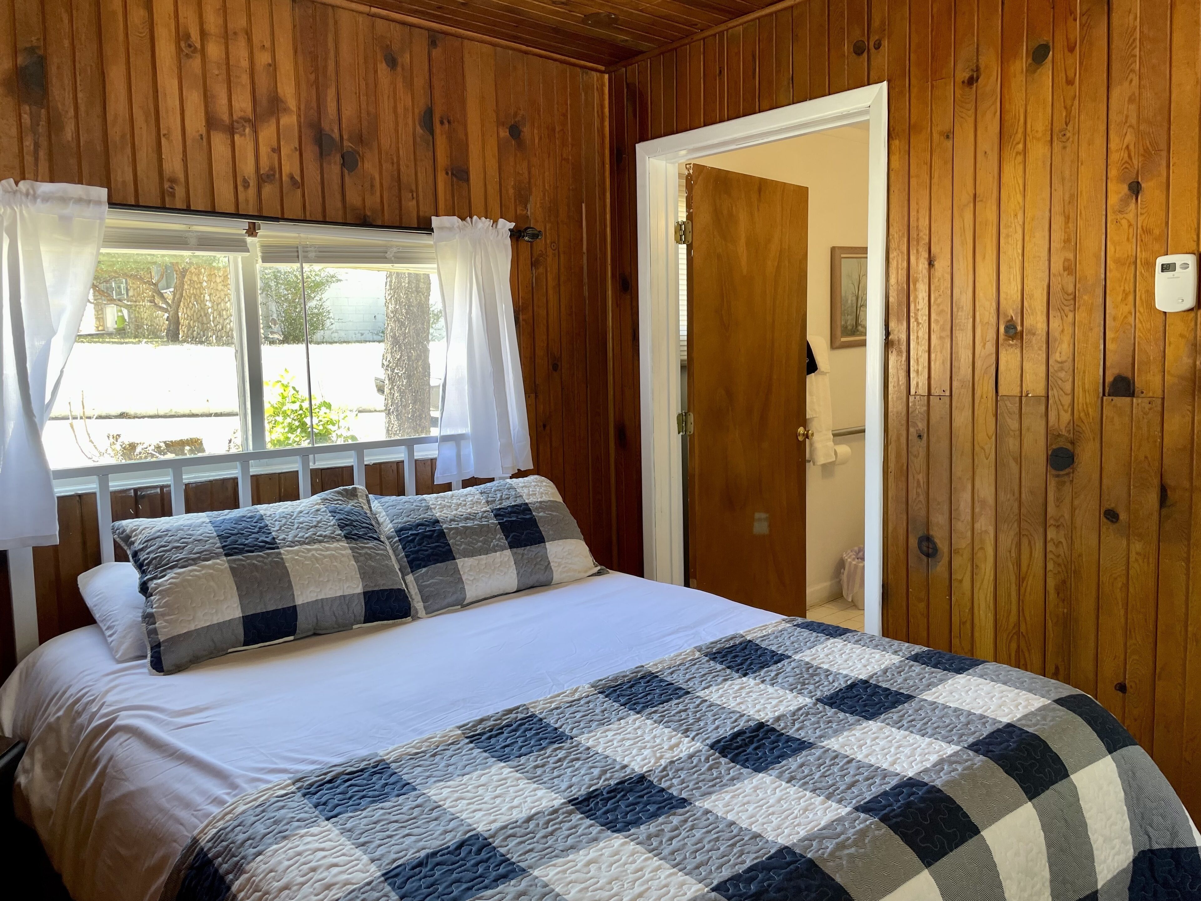 Une pièce aux murs lambrés, avec un lit, une fenêtre avec des rideaux et une porte donnant sur une salle de bain.