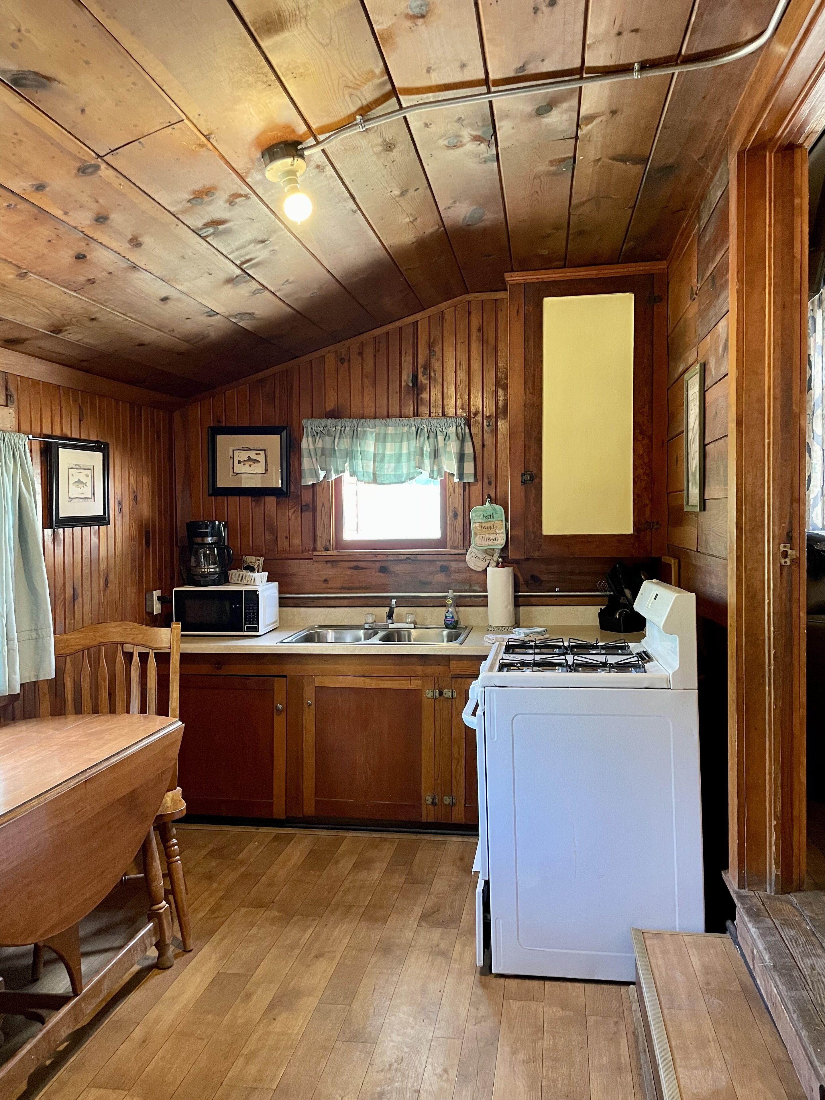 A cabin-style kitchen with wooden walls, a window with a plaid valance, a sink, a stove, and a microwave.