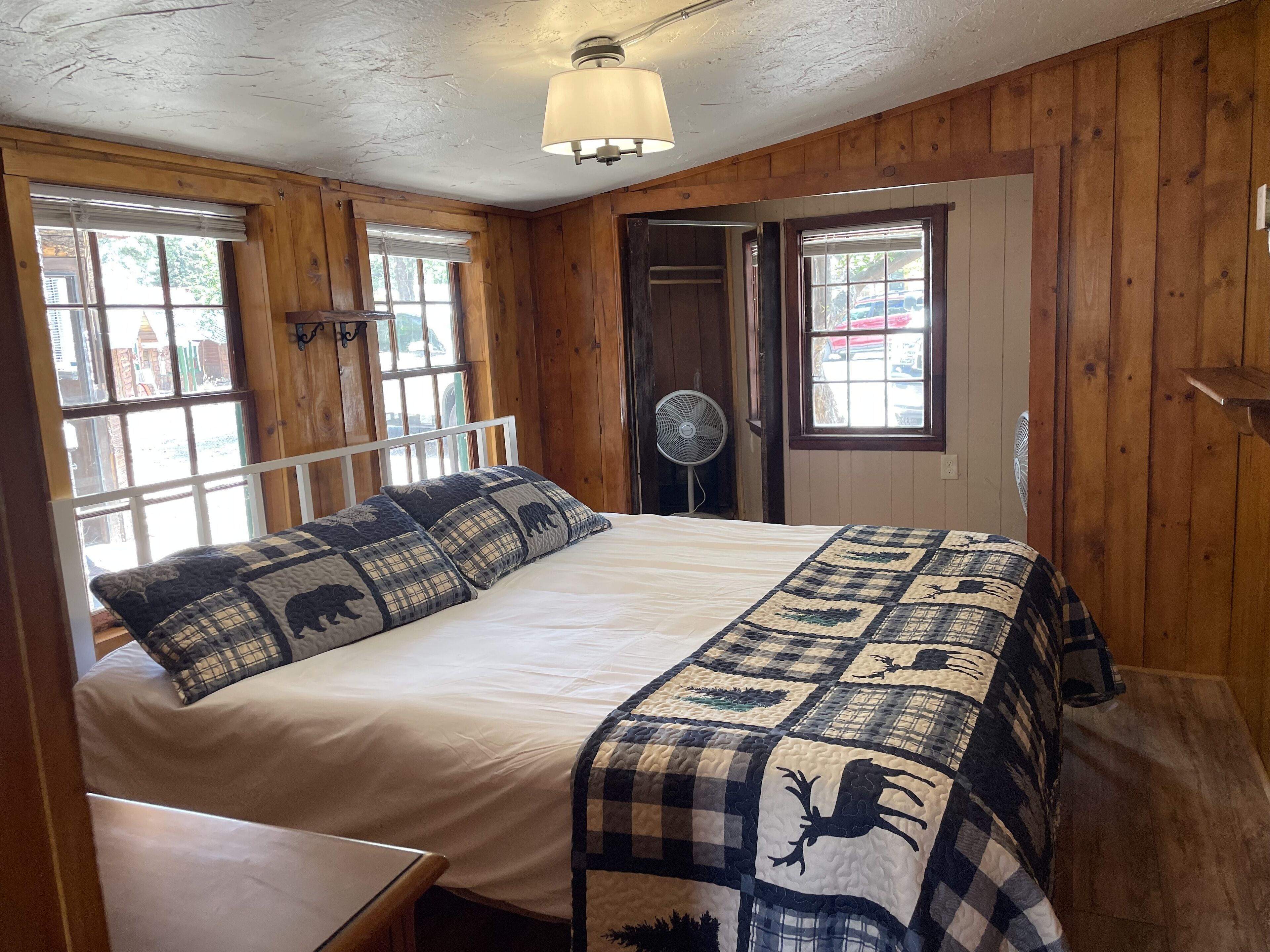 A wooden-paneled room with a bed, two windows, a ceiling fan, and a light fixture.