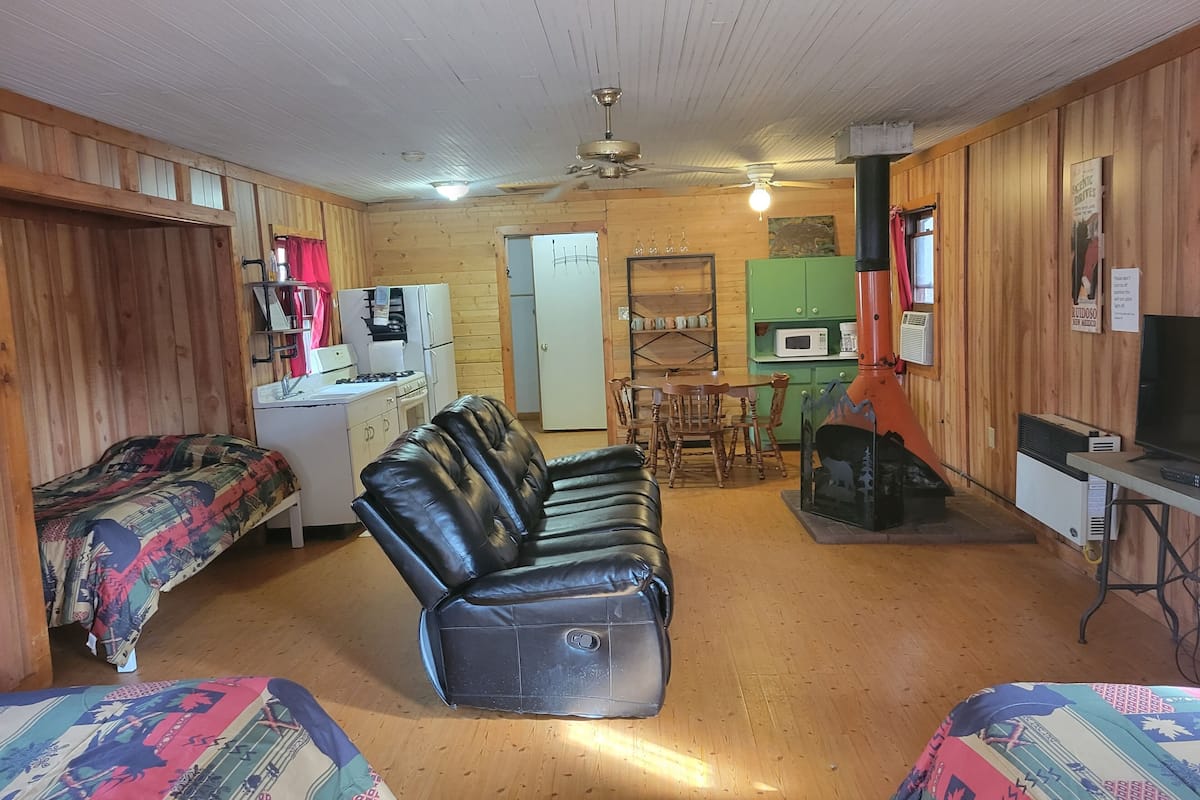 A cozy cabin-style room with a bed, a sofa, a dining table with chairs, a kitchenette, and a fireplace.