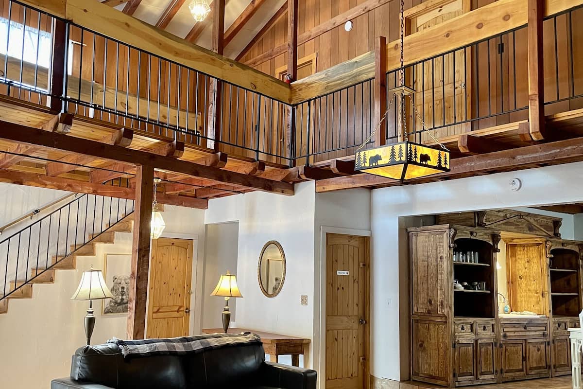 A spacious loft-style living area with a high ceiling, exposed wooden beams, a black leather sofa, a wooden coffee table, and a rustic kitchen area with wooden cabinets.
