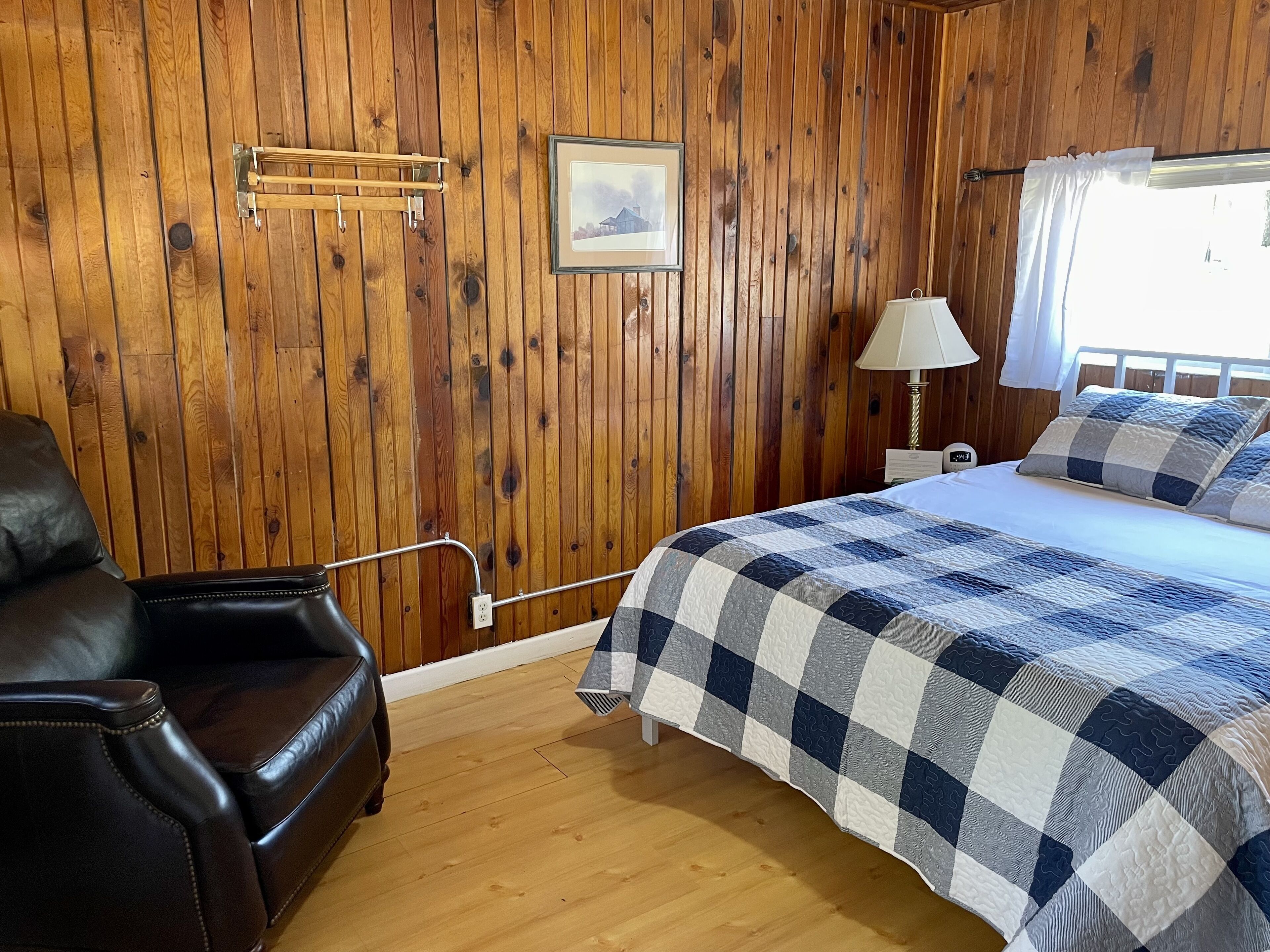 A wooden-paneled room with a bed, a leather chair, a bedside lamp, and a framed picture on the wall.