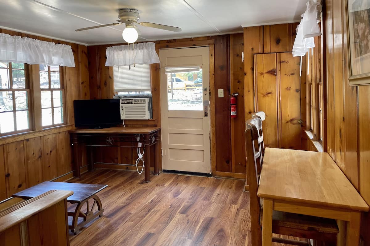 A compact room with wooden walls and floors, a ceiling fan, a television, a small table, and a window with curtains.