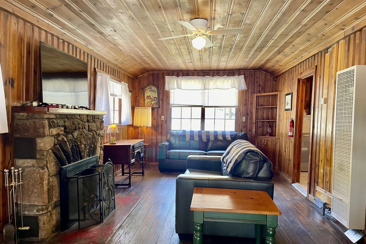 A cozy cabin interior with a stone fireplace, wooden walls, a blue sofa, and a wooden table.