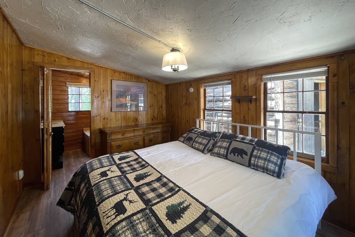 A wooden cabin bedroom with a bed, dresser, and windows.