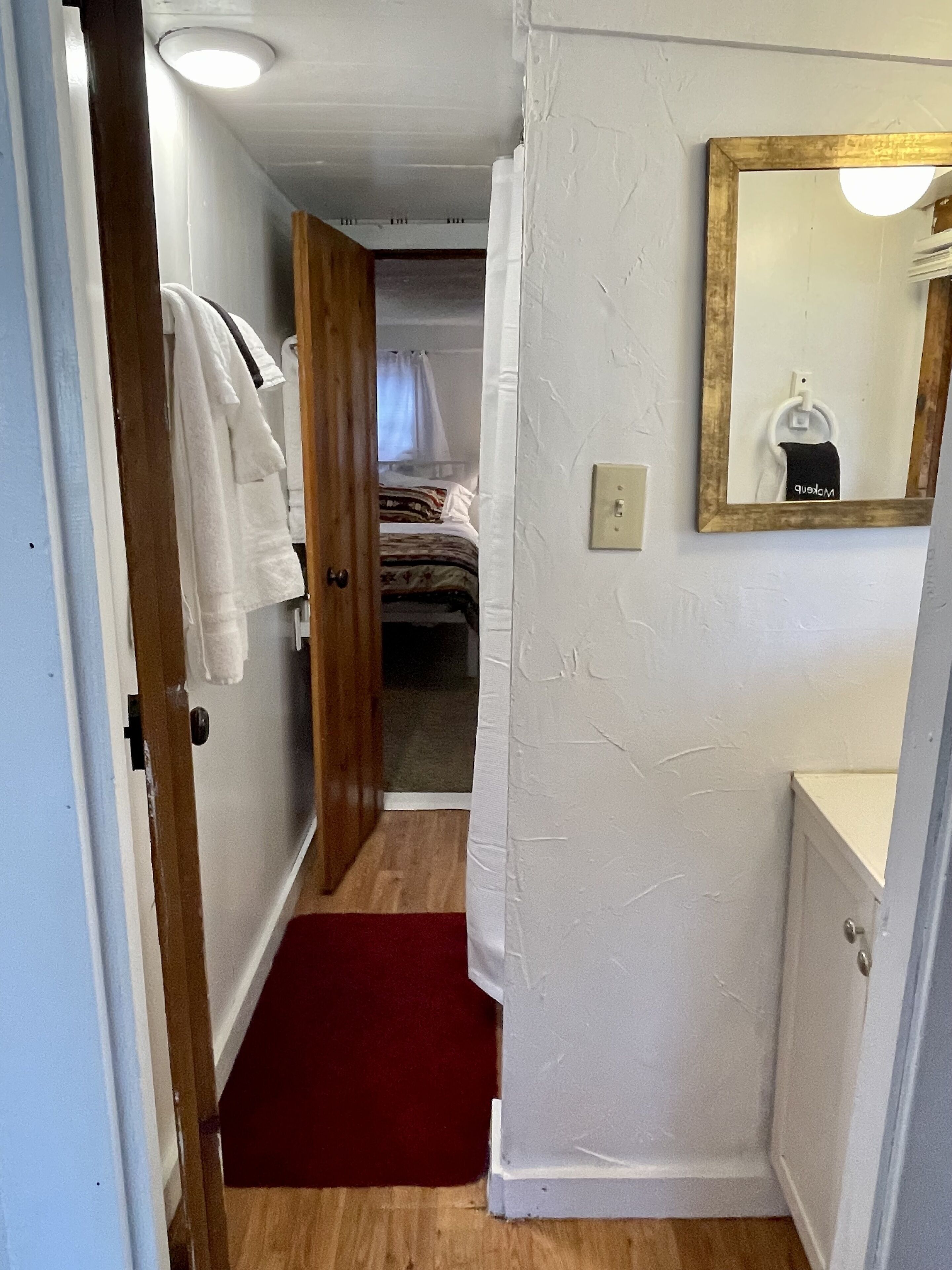 A narrow hallway with a wooden door leading to a bedroom with a bed and a white curtain. A red rug is on the floor, and a mirror with a gold frame hangs on the wall.