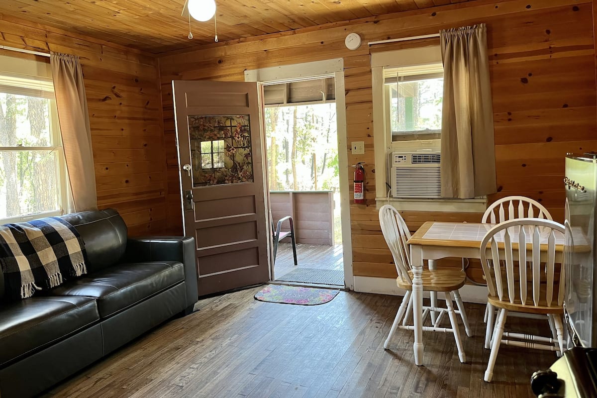 A cozy cabin interior with a wooden ceiling, a ceiling fan, a black leather sofa, a dining table with white chairs, and a window with curtains.