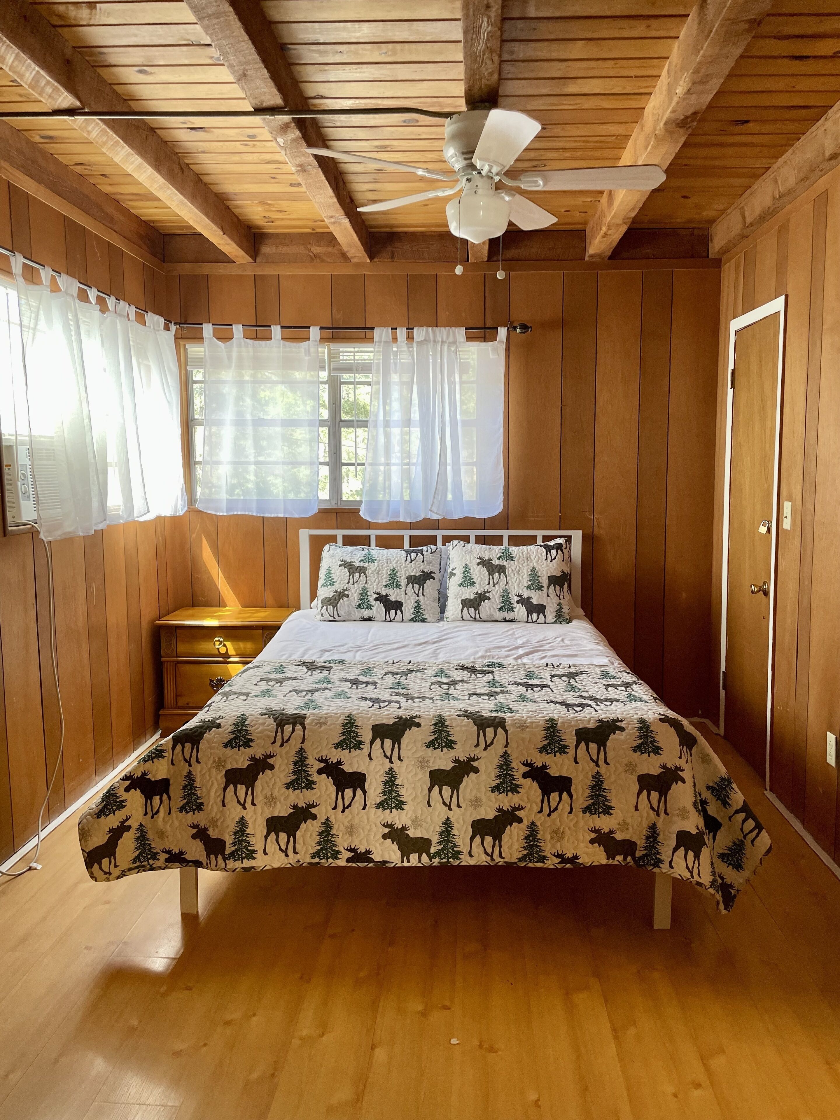 Un dormitorio con techo de madera, una cama con edredón de temática de alce, una mesita de noche y una ventana con cortinas traslúcidas.