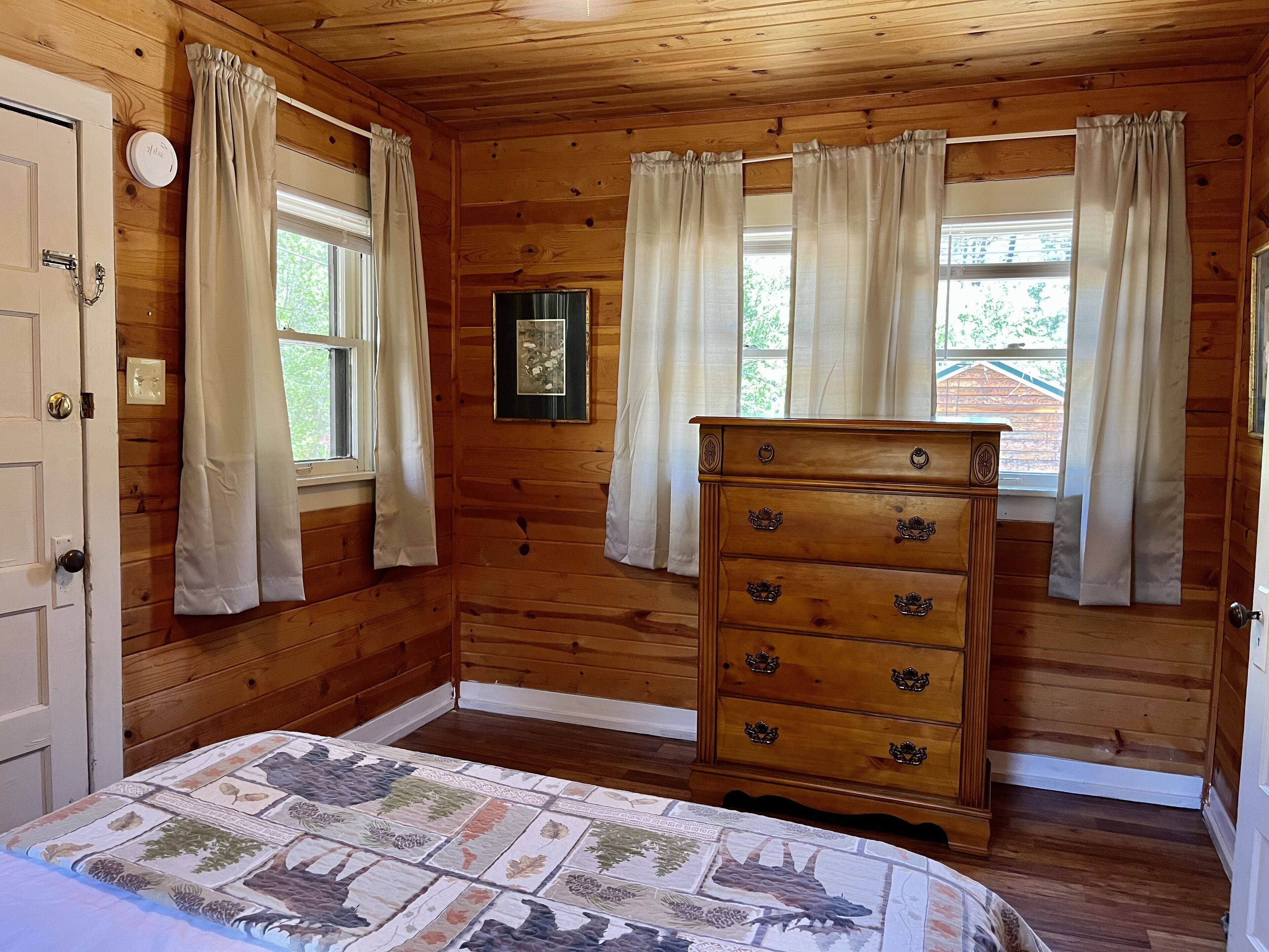 Une chambre dans une cabane en bois, avec un lit, une commode et deux fenêtres équipées de rideaux.