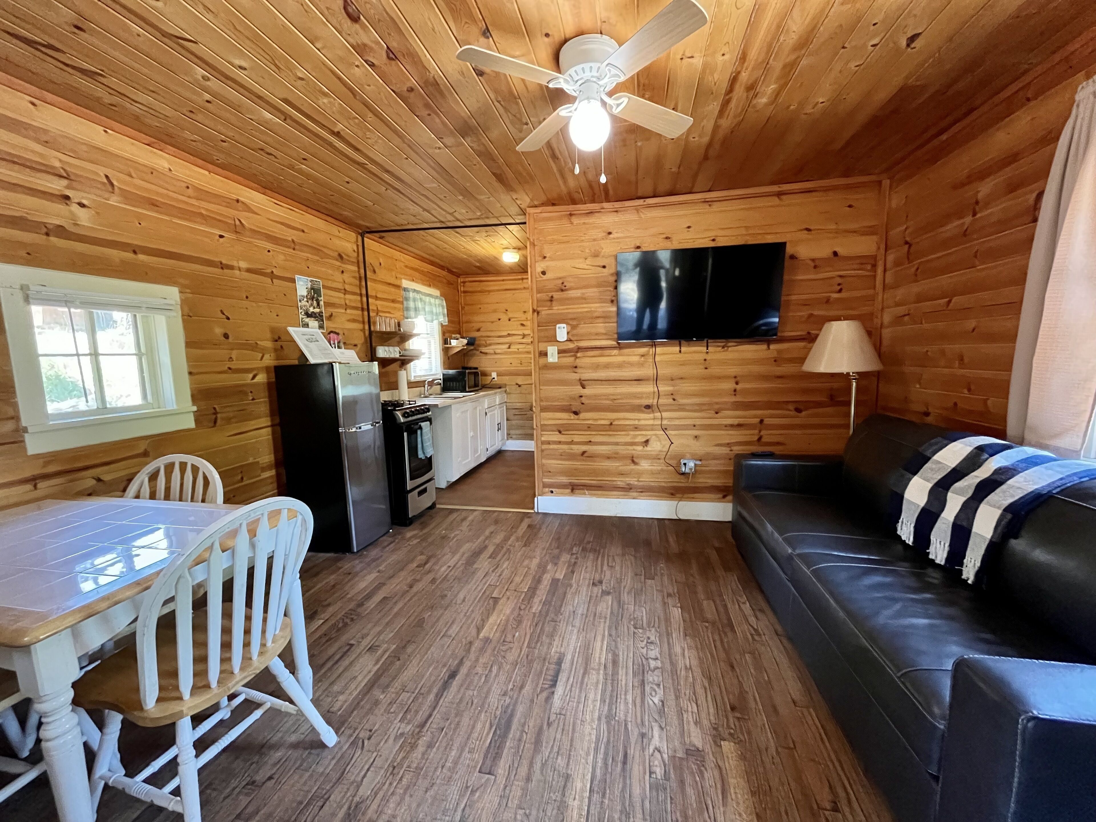 A cozy cabin interior with a dining table, chairs, a sofa, a TV mounted on the wall, a ceiling fan, and a kitchen area with a refrigerator and microwave.