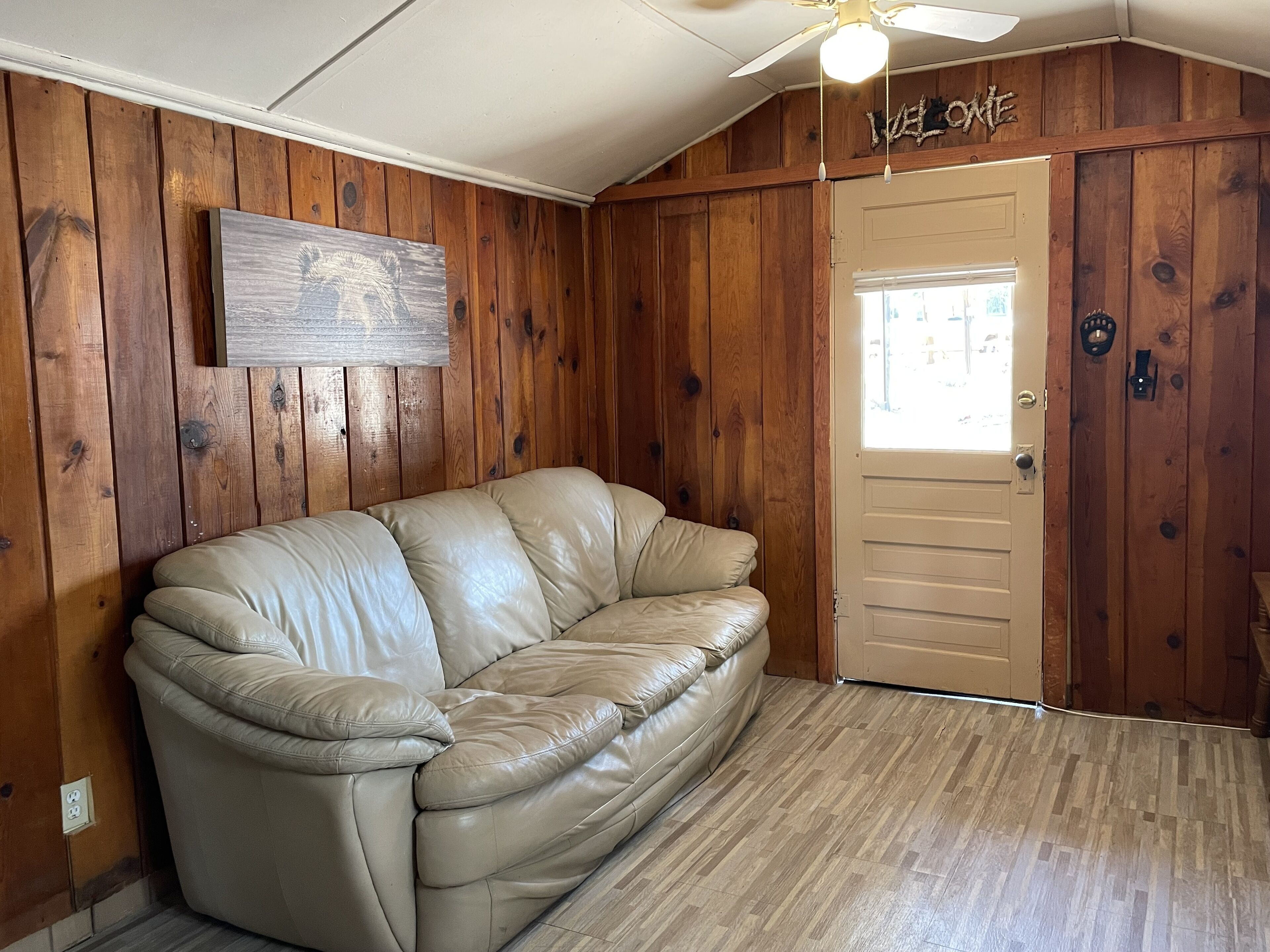 A room with a beige leather sofa, wooden walls, a ceiling fan, and a door with a 'Welcome' sign.