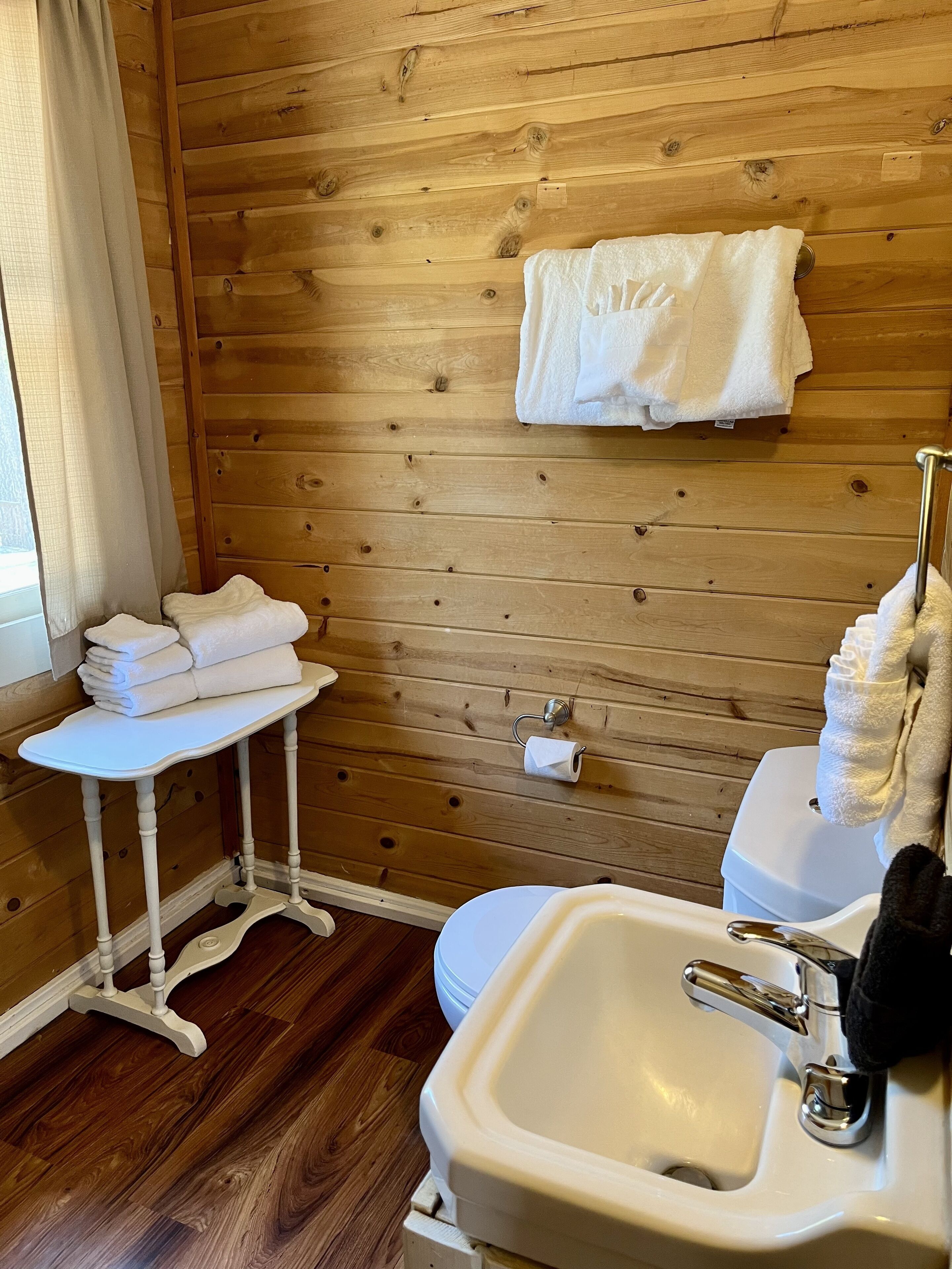 A wooden-paneled bathroom with a white sink, toilet, and a small table with stacked towels.