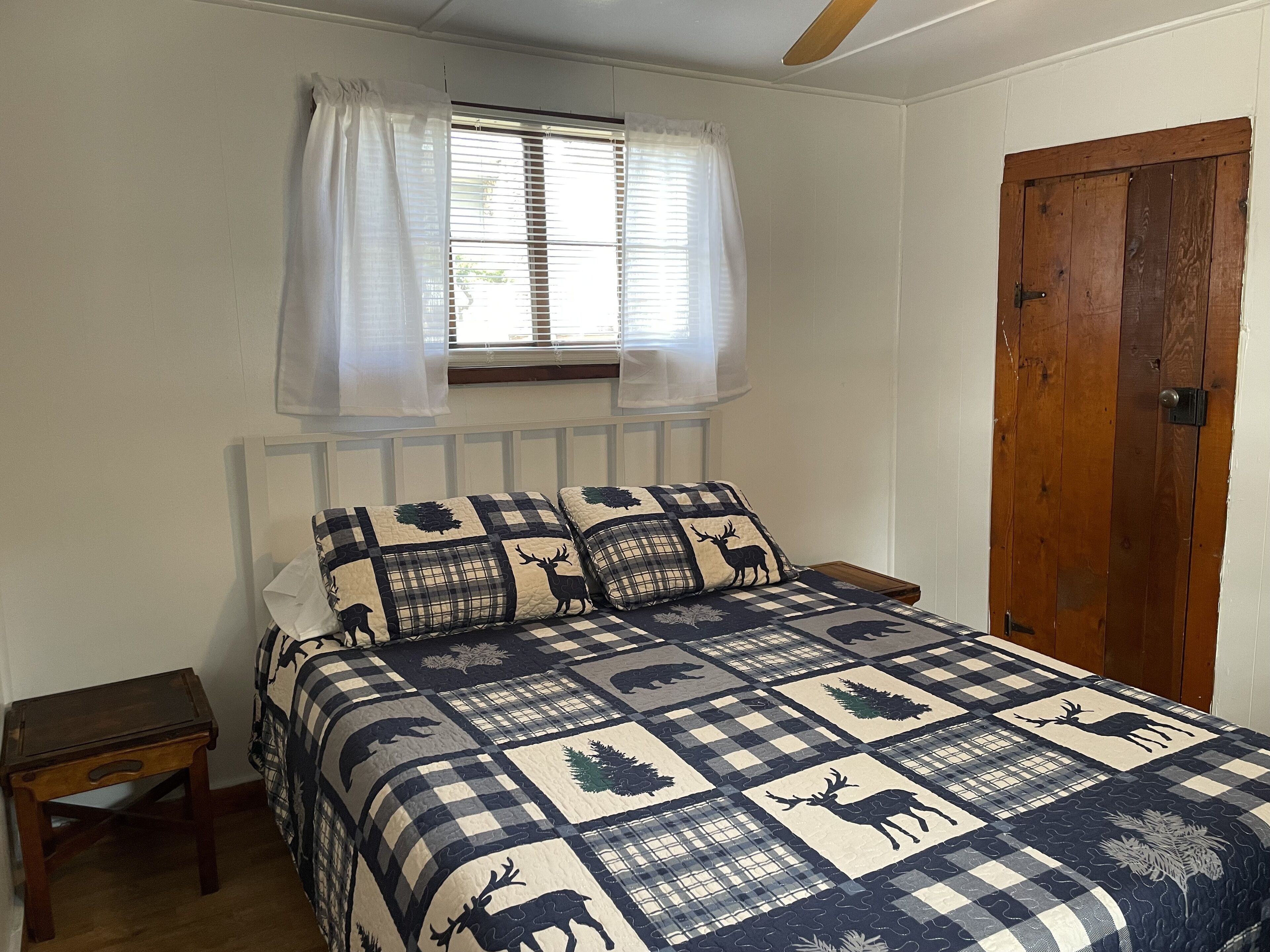 A bedroom with a checkered bedspread featuring animal prints, a wooden door, and a window with sheer curtains.