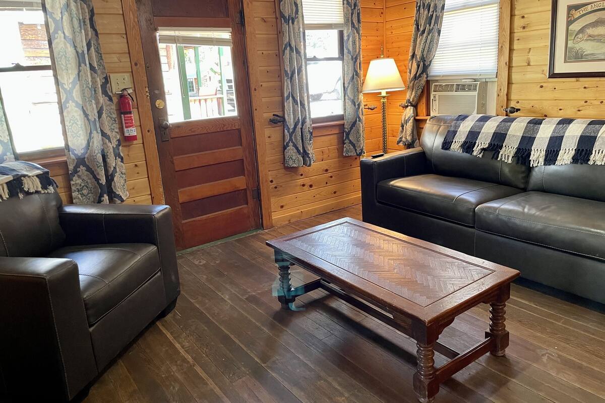 A cozy cabin interior with a wooden door, a leather sofa, a wooden coffee table, and a lamp.