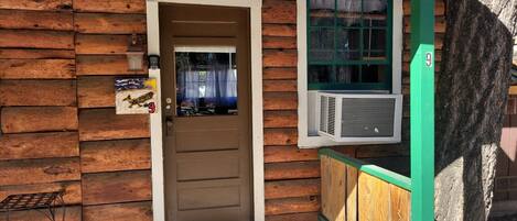 A wooden cabin with a brown door, a window with a green frame, and an air conditioning unit mounted on the wall.