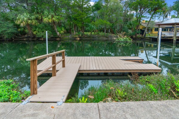A wooden dock extending into a calm water body, surrounded by lush greenery and a small structure in the background.
