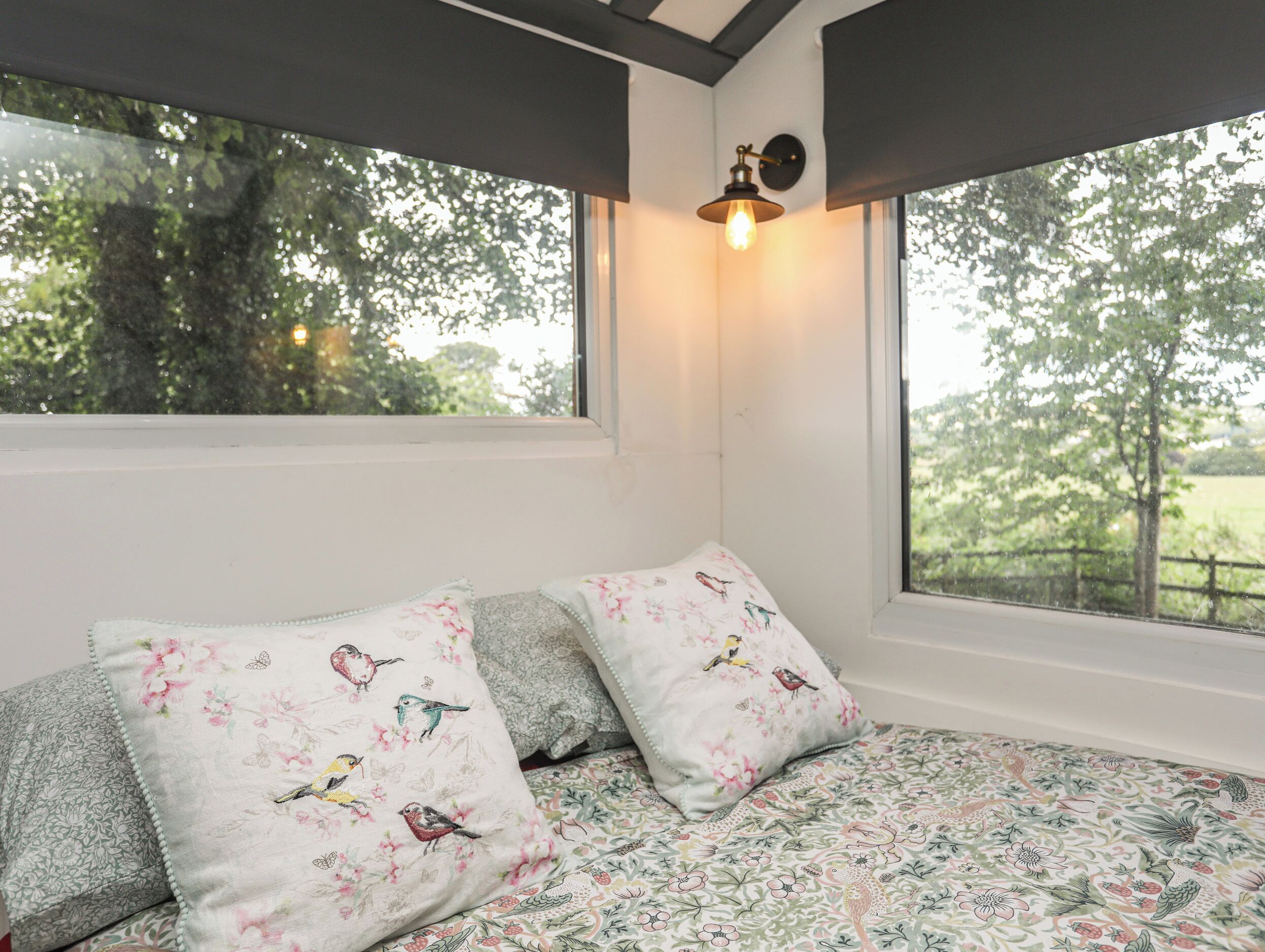 Een slaapkamer met een groot raam, beddengoed met bloemenprint en twee kussens met vogelmotieven.