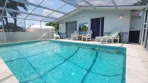 Une piscine à l’eau d’un bleu cristallin, entourée d’un patio abrité avec des chaises longues et d’un bâtiment doté de grandes fenêtres.