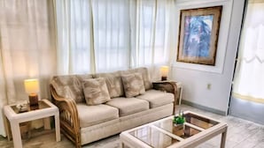 Un salon avec un canapé beige, deux tables d’appoint avec des lampes, une table basse en verre et un tableau encadré au mur.