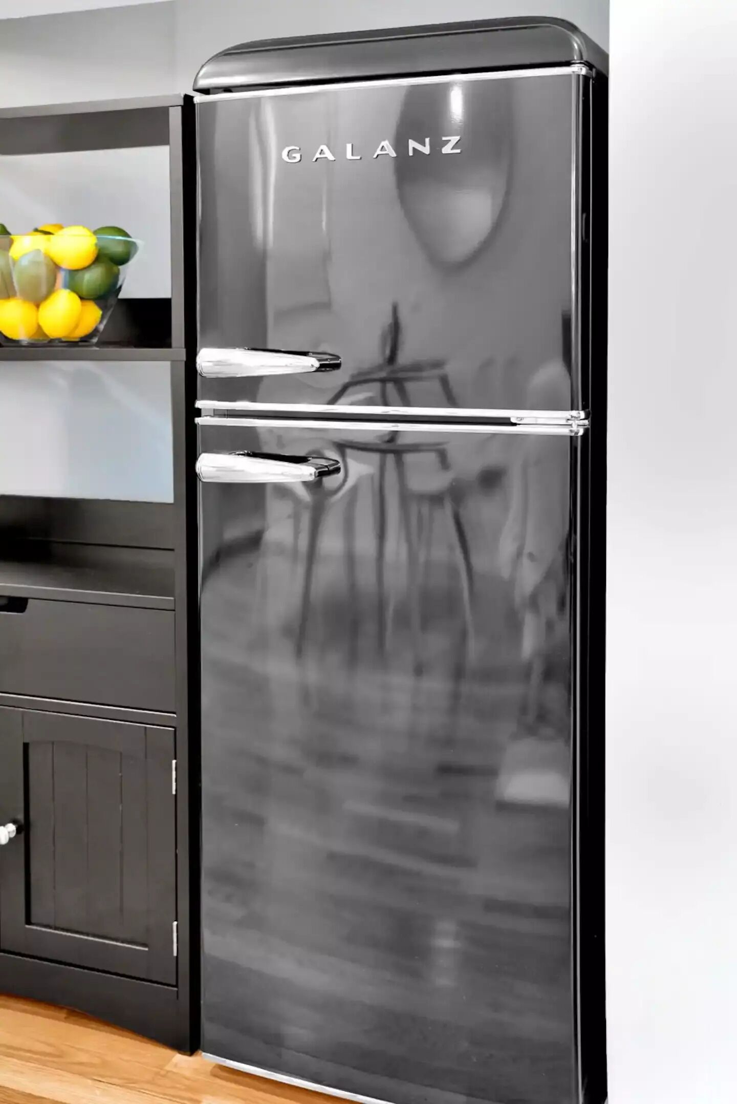 A black Galanz refrigerator with a reflective surface, placed in a kitchen with a wooden cabinet and a bowl of lemons on the counter.