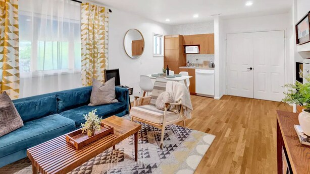 Una sala moderna con un sofá azul, una mesa de centro de madera y un tapete estampado. Hay una zona de cocina con armarios blancos y un microondas.