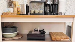 Una cocina con cafetera, hervidor y tostadora sobre la encimera. Hay ollas y una tabla de cortar en los estantes inferiores.