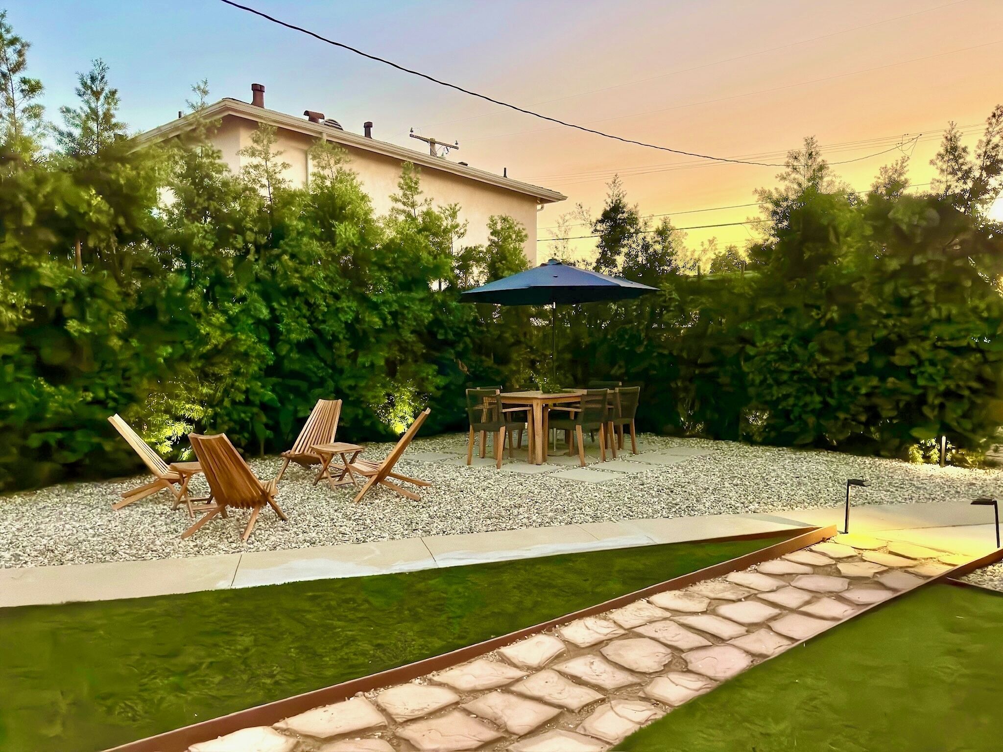 A backyard with a patio table and chairs, surrounded by a gravel area and a small water feature.
