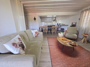 Ein Wohnzimmer mit einer Couch, einem Couchtisch und einem Sessel. Im Hintergrund ist eine Küche mit einem Esstisch und Stühlen zu sehen.