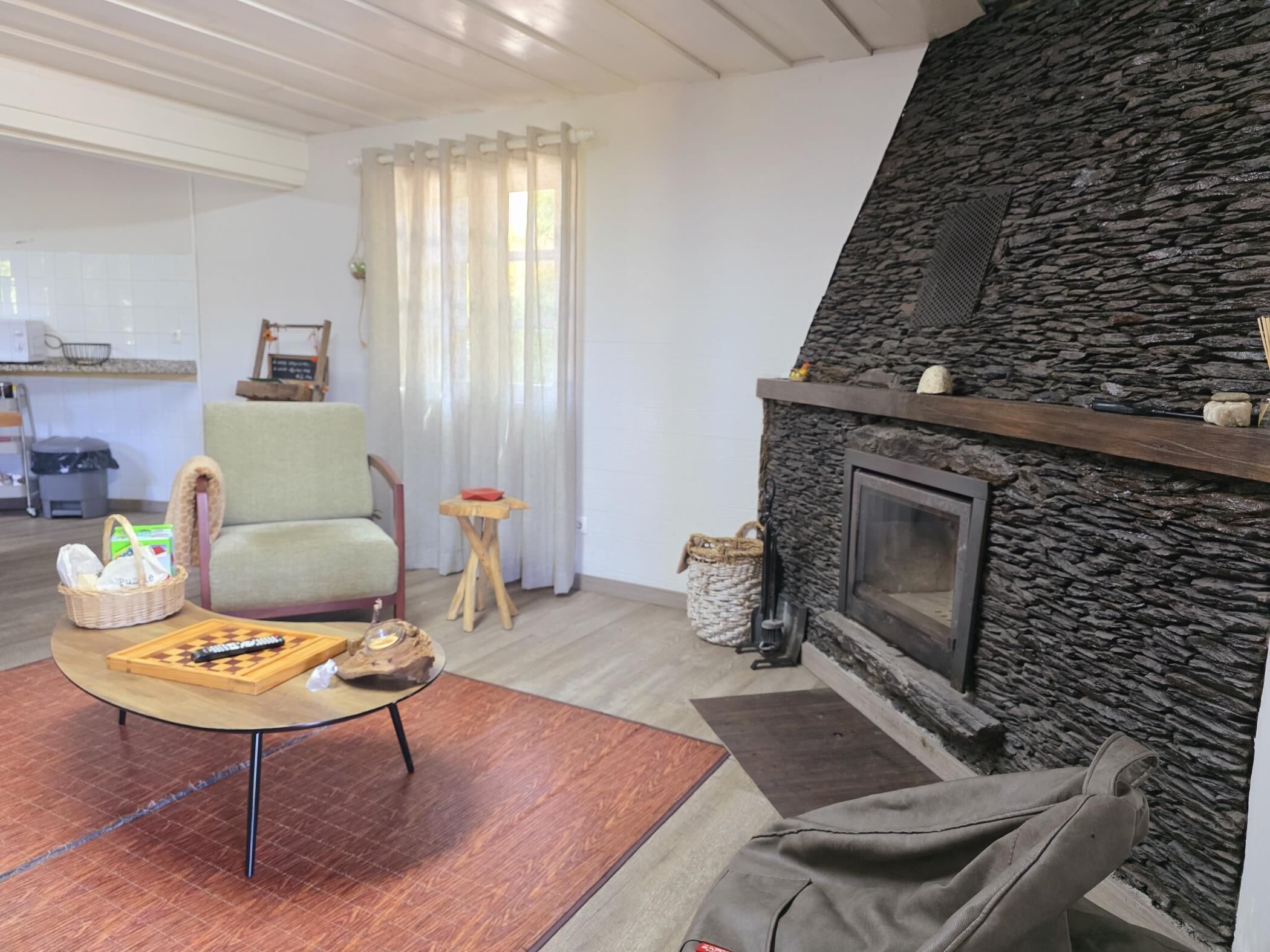 Een woonkamer met een stenen openhaard, een groene fauteuil, een salontafel met een puzzel, en een keuken op de achtergrond.