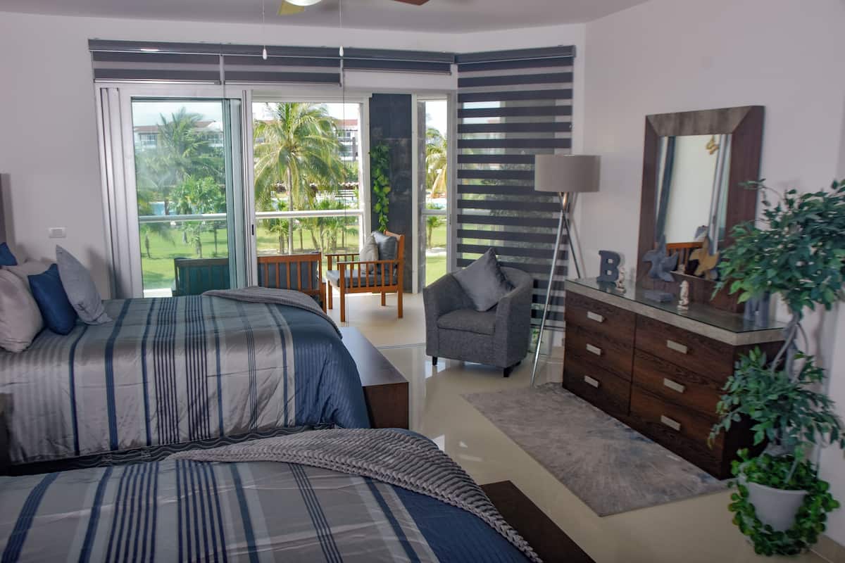 Ein Schlafzimmer mit Bett, Stuhl, Schminktisch und einem Balkon mit Blick auf Palmen.
