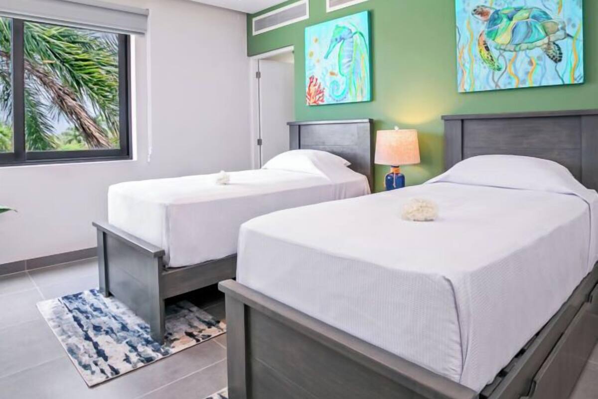 Ein Hotelzimmer mit zwei Einzelbetten, einem Deckenventilator, einem Fenster mit Blick auf Palmen und Kunstwerken an den Wänden.