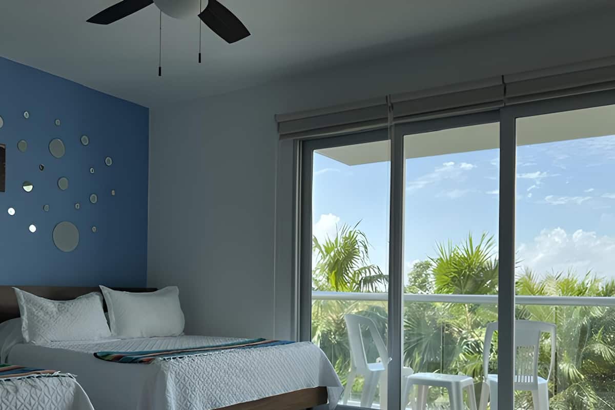 Ein Schlafzimmer mit Bett, Deckenventilator, großem Fenster mit Blick auf Palmen und Wanddekoration.
