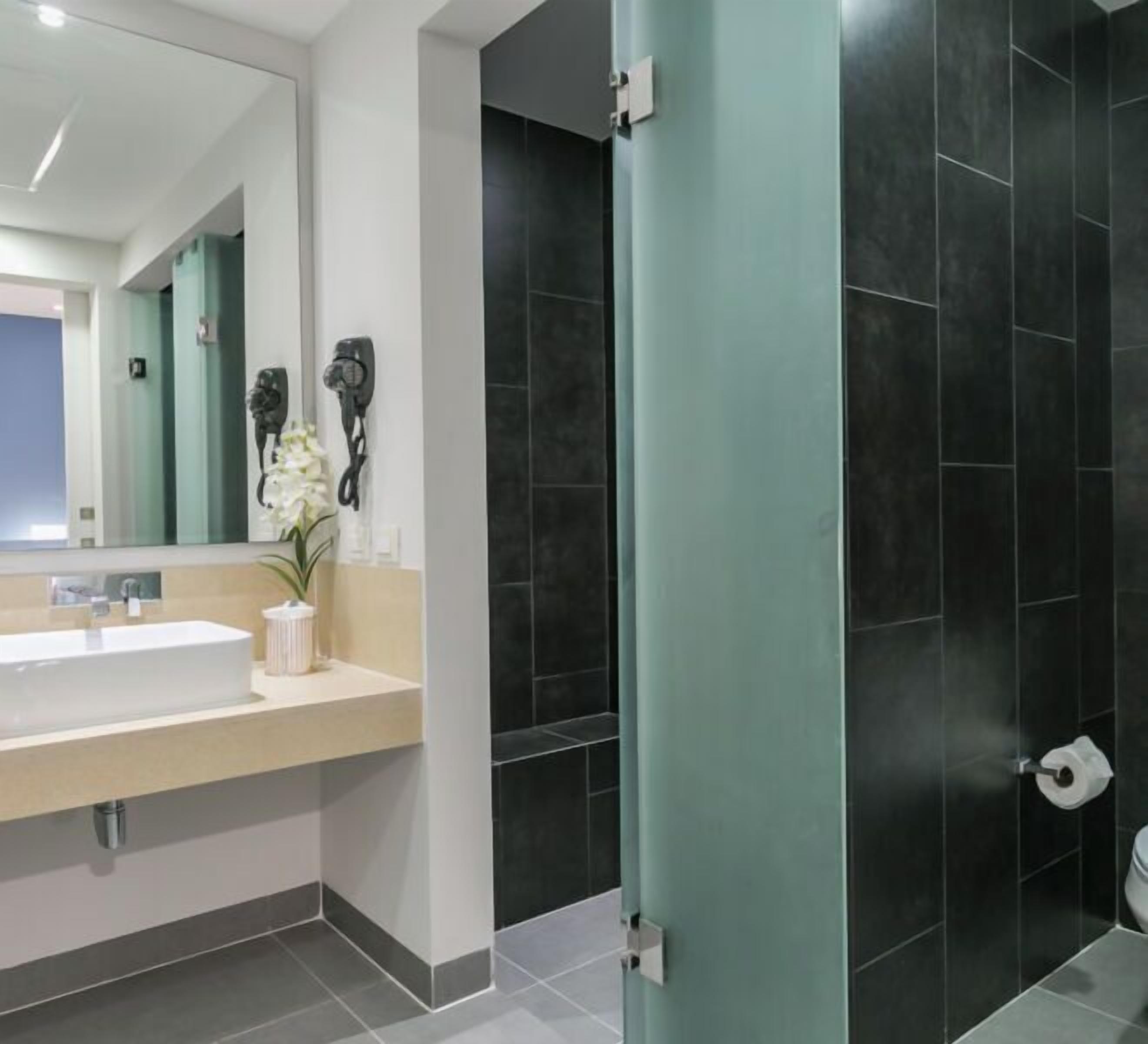 A modern bathroom with a large mirror, a white sink, a hairdryer, and dark tiled walls.