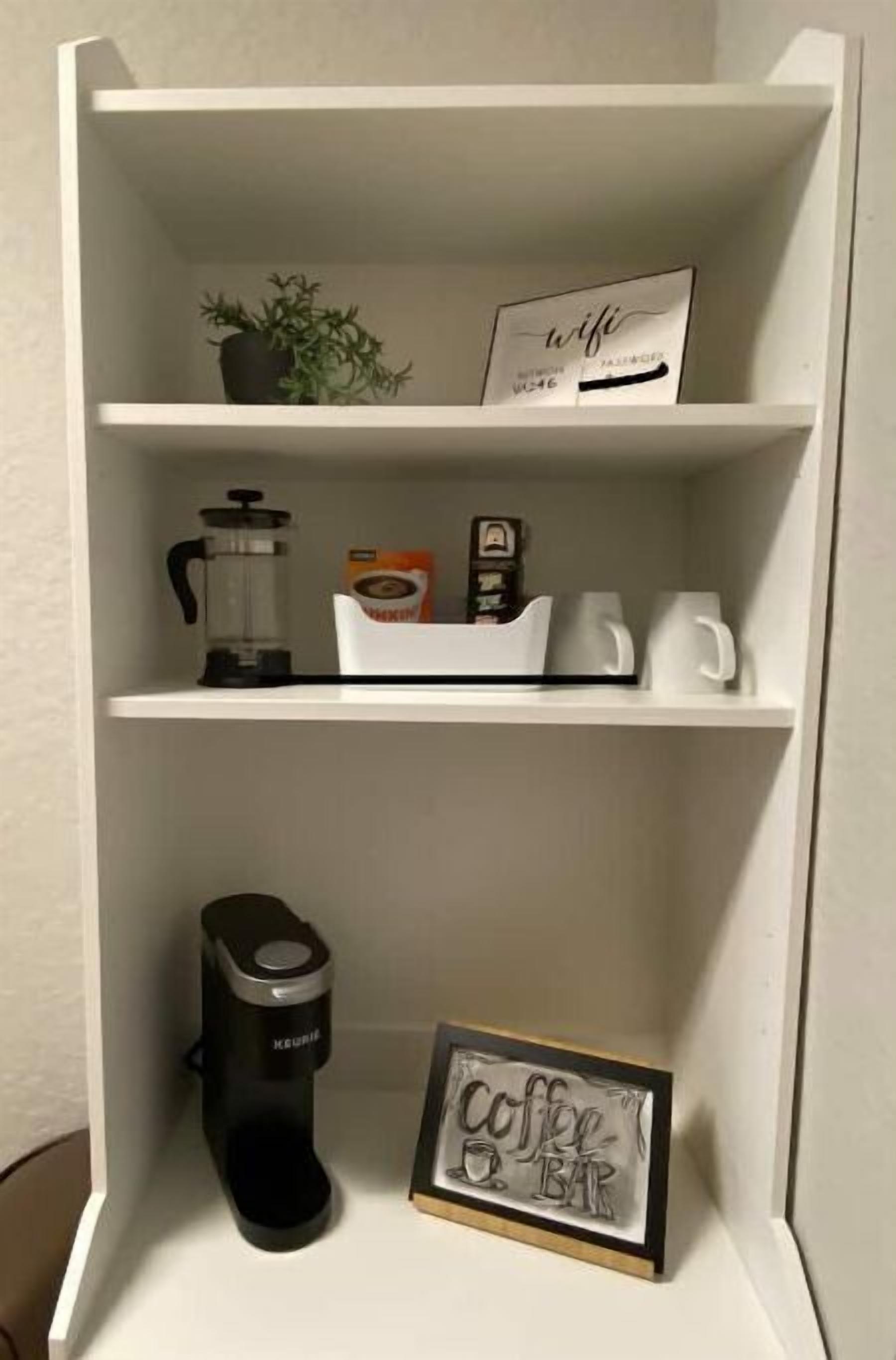 Une étagère blanche avec une machine à café, une enseigne indiquant « bar à café » et une plante en pot.