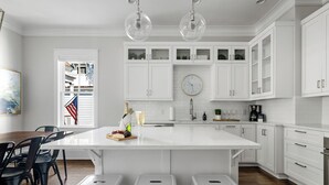 A modern kitchen with white cabinetry, a large island with stools, a clock, and a view of a balcony with an American flag. - Old Village | Amazing Location | The Big Spoon (Mt Pleasant)