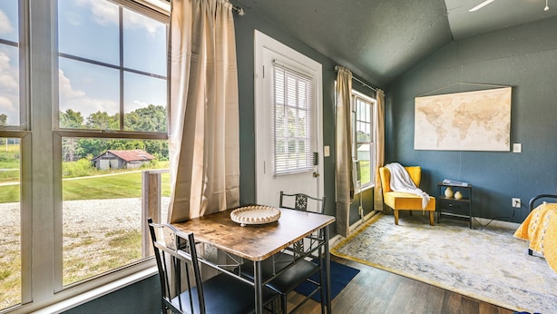 Een kamer met een groot raam, een eettafel met stoelen, een bed met een gele deken, een stoel met een kussen, een kleine tafel met een wereldbol, en een kaart aan de muur.