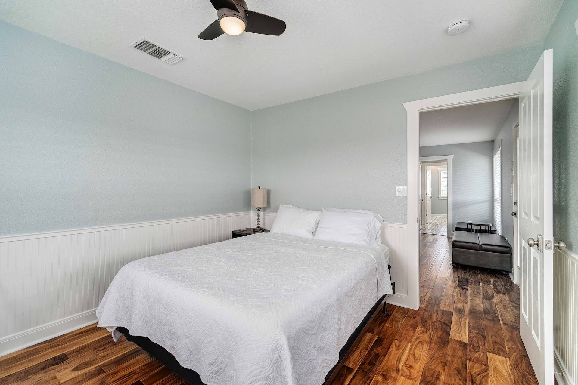 A bedroom with a bed, a nightstand, a lamp, a ceiling fan, and a door leading to another room.