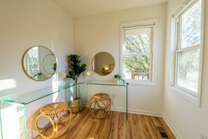 Bathroom features a glass vanity, round mirror, and a potted plant, with a wicker stool and large window.