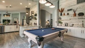 Een ruime woonkamer met een pooltafel, moderne verlichtingsarmaturen en een keuken voorzien van witte kastjes en decoratieve elementen.