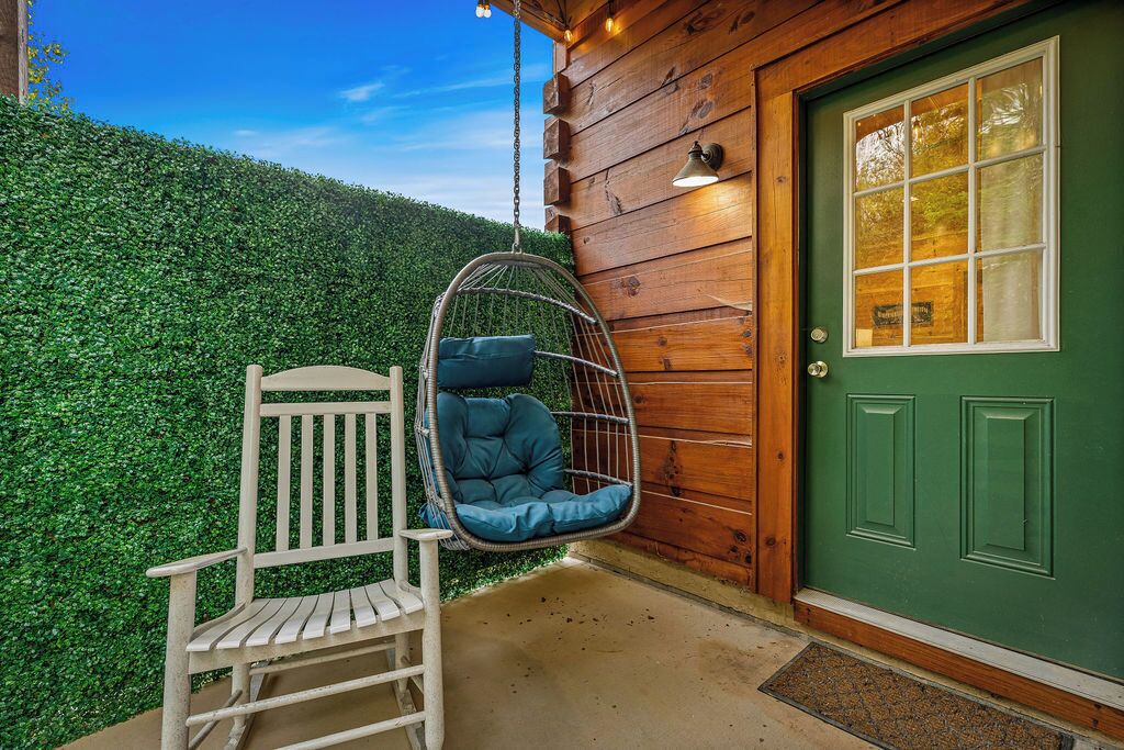 Pared exterior de madera, una puerta verde, un banco blanco y una silla colgante con cojines.