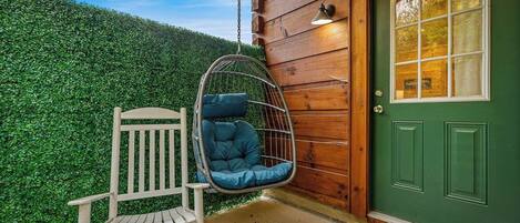 Pared exterior de madera, una puerta verde, un banco blanco y una silla colgante con cojines.