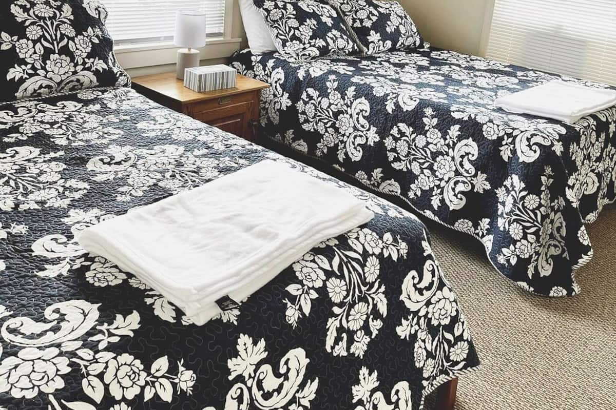 A room with two beds, each with a floral-patterned bedspread, a bedside table with a lamp, and a window with blinds.