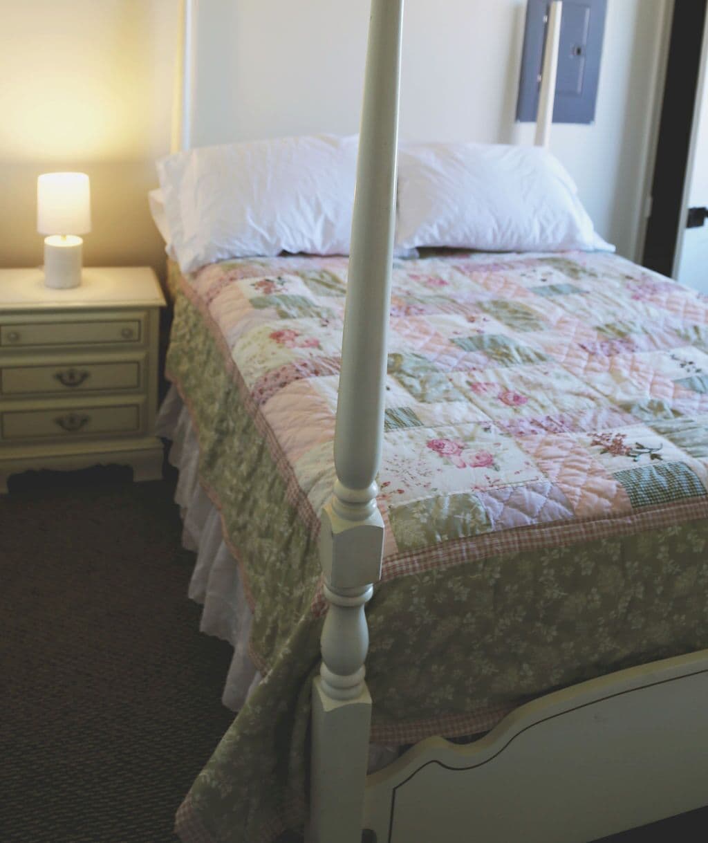 Una cama con dosel, colcha estampada, mesita de noche con lámpara e interruptor de luz en la pared.
