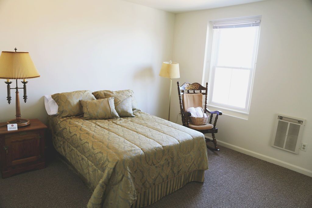 A neatly made bed with a patterned bedspread, two bedside lamps, a chair with a cushion, and a window with blinds.